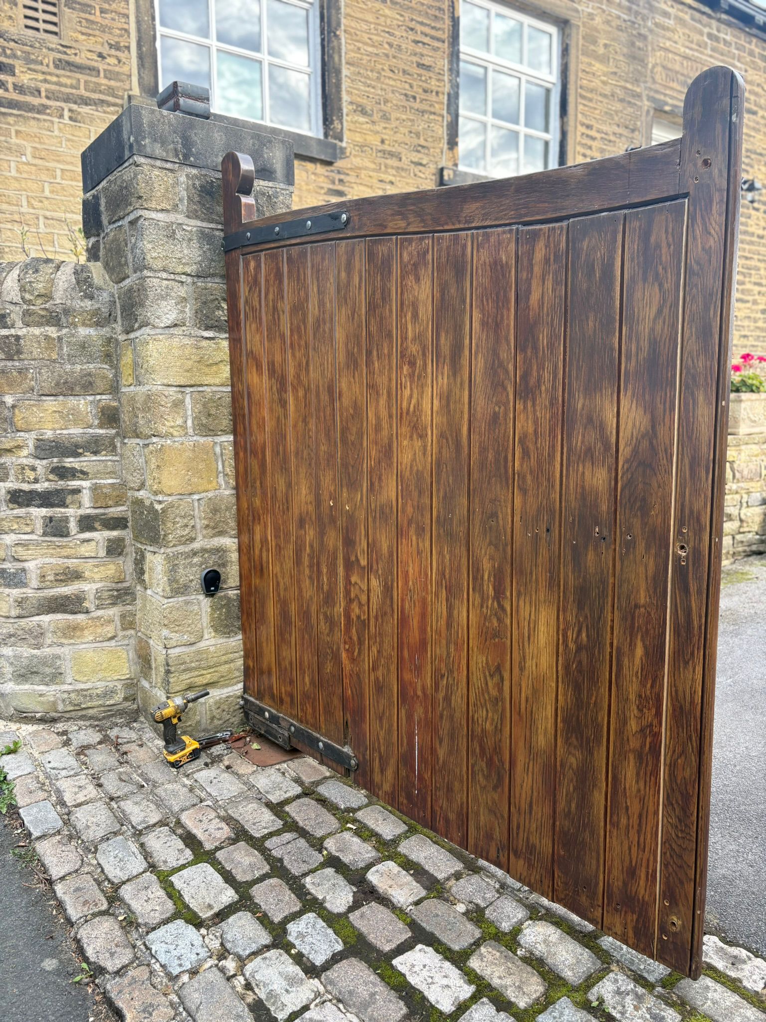 A set of large, dark-stained wooden driveway gates attached to a stone pillar, showing the initial stage of an automation retrofit with a yellow power drill on the cobblestone ground.