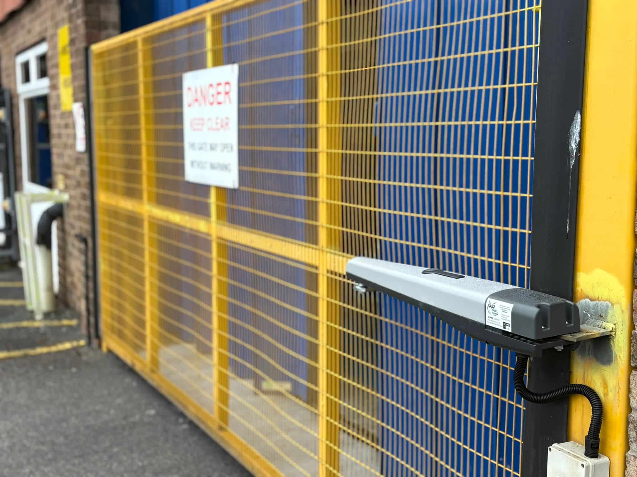 A detailed view of a Nice Toona automation arm fixed to a heavy-duty yellow mesh gate. The image captures the sturdy mounting bracket and the protective conduit for the wiring, showcasing the robust engineering required for industrial access control.