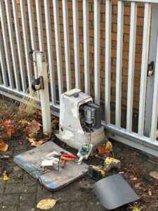 An automated sliding gate motor with its protective casing removed, showing internal wiring during a repair by Owl Access Ltd. Tools including a power drill, pliers, and a multimeter are laid out on a service mat on the ground. In the background, a white metal security gate and infrared safety sensors are visible.