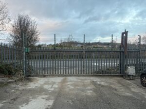 A large industrial sliding cantilever gate made of silver steel palisade fencing. The gate features integrated electric security wiring along the top and is set against a backdrop of a paved driveway, a public road, and a residential hillside under a cloudy sky.