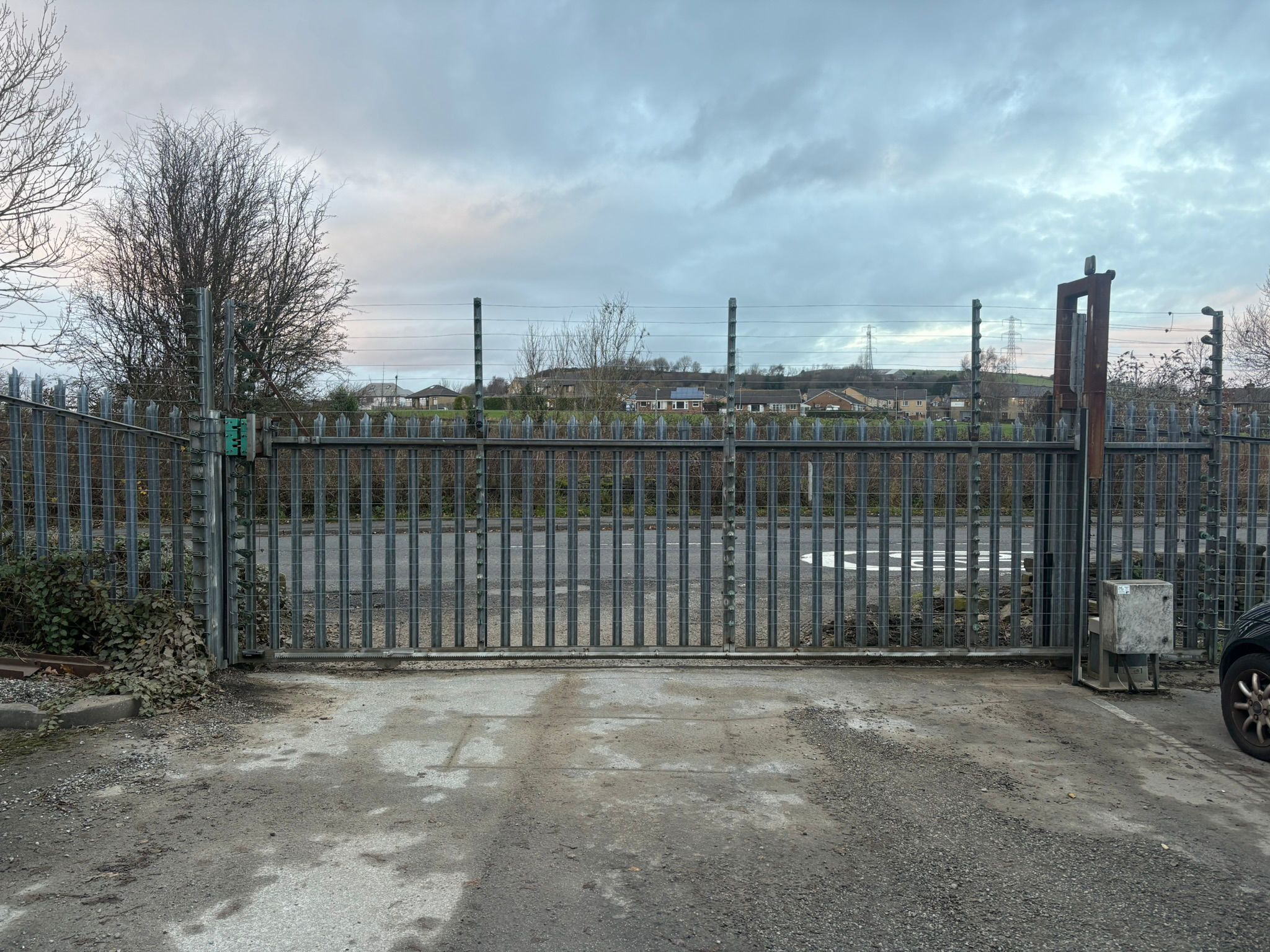 A large industrial sliding cantilever gate made of silver steel palisade fencing. The gate features integrated electric security wiring along the top and is set against a backdrop of a paved driveway, a public road, and a residential hillside under a cloudy sky.