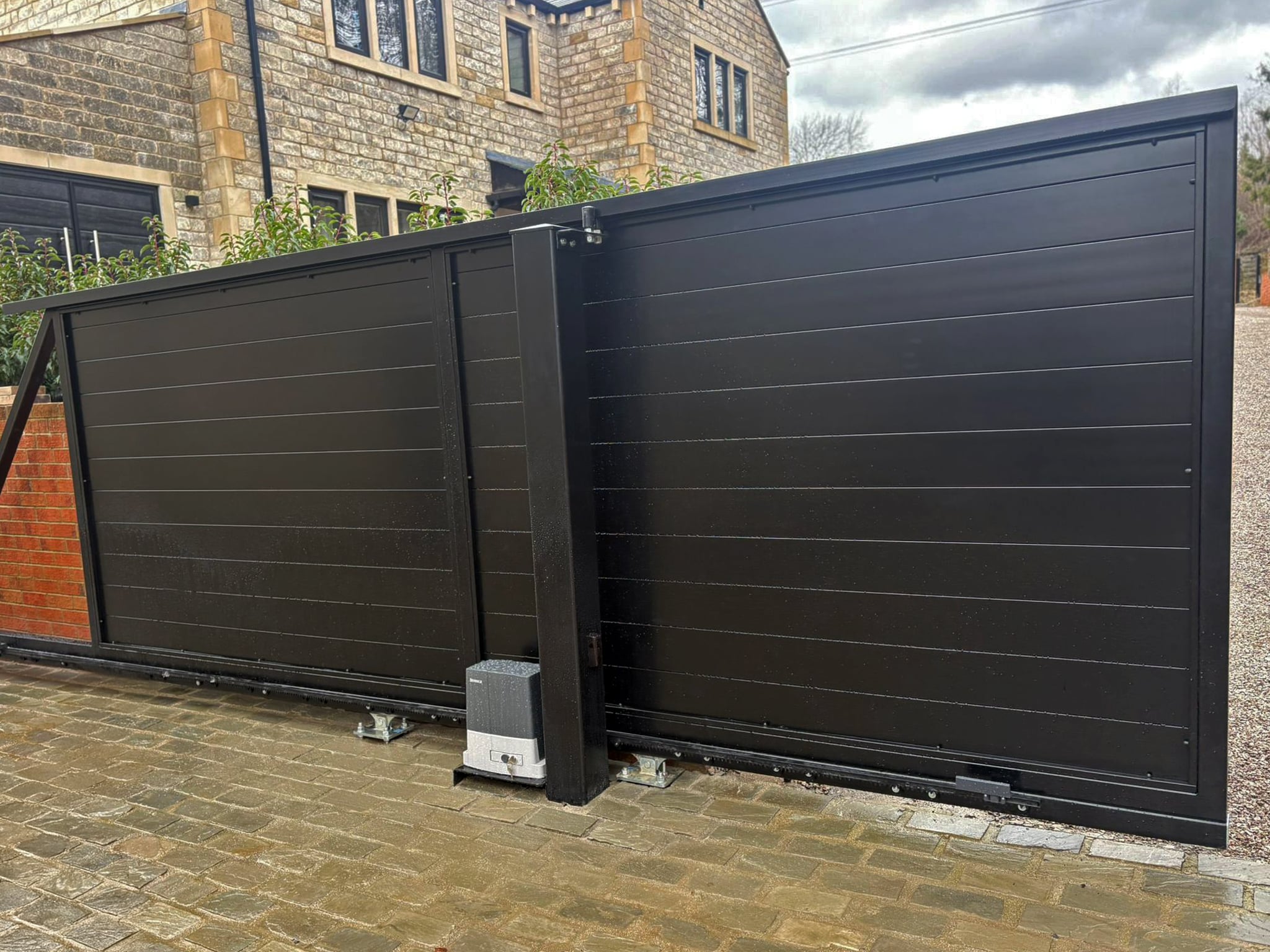 A side profile view of a black sliding gate in the partially open position, highlighting the Beninca automation motor and the sleek horizontal slat design.