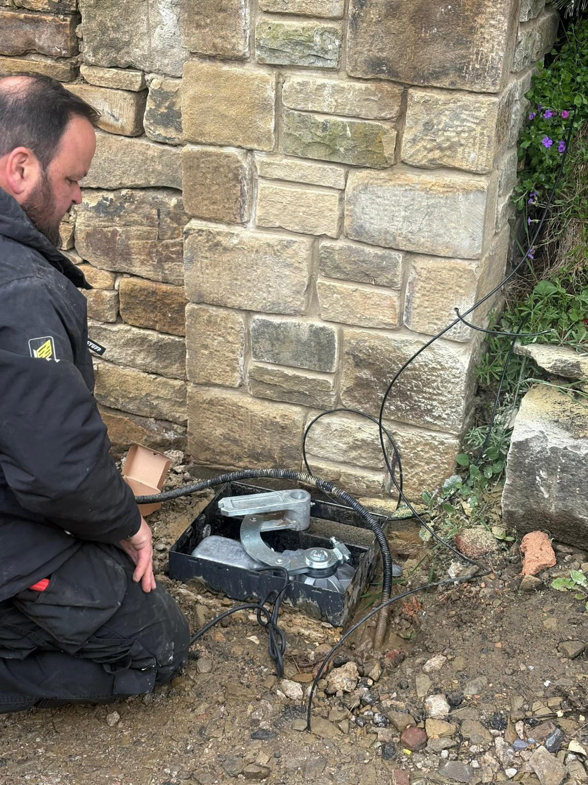 An engineer in a black work jacket is kneeling on the ground next to a stone pillar, installing a Nice underground gate automation motor inside its foundation box.