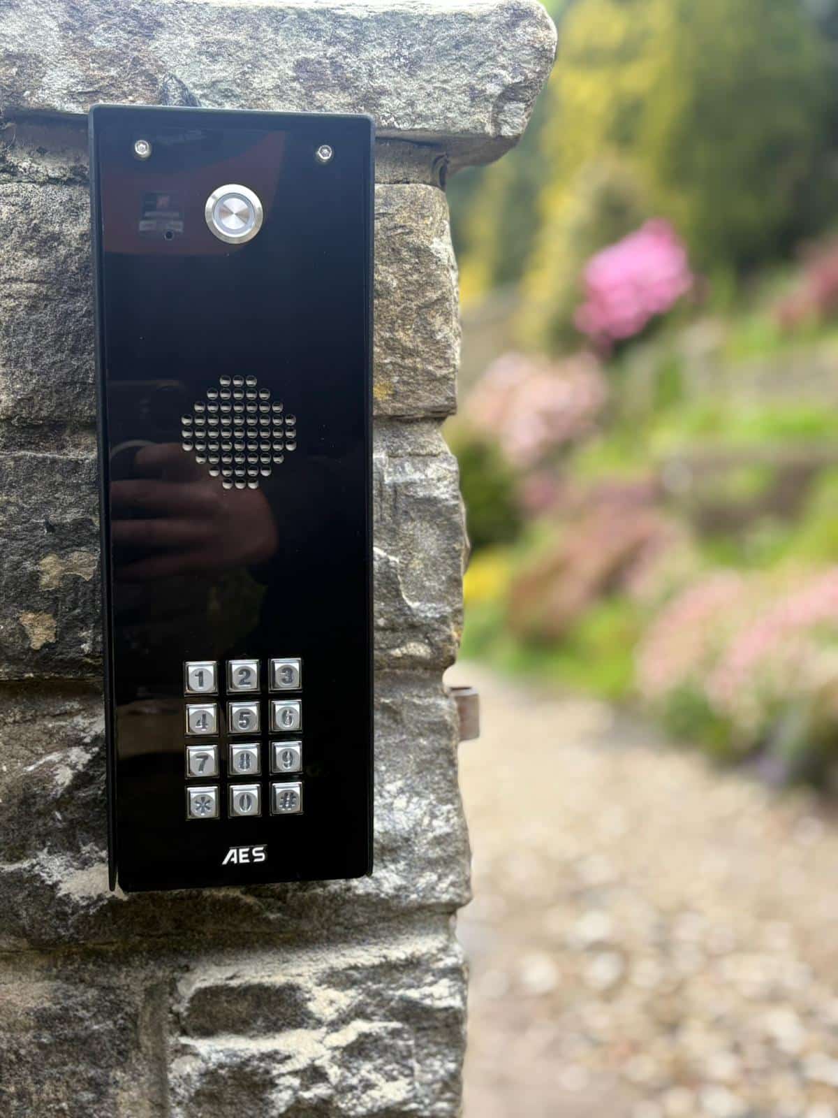 A close-up of a sleek, black AES Global intercom panel with a metallic keypad and speaker grill, mounted onto a stone entrance pillar with a blurred garden background.