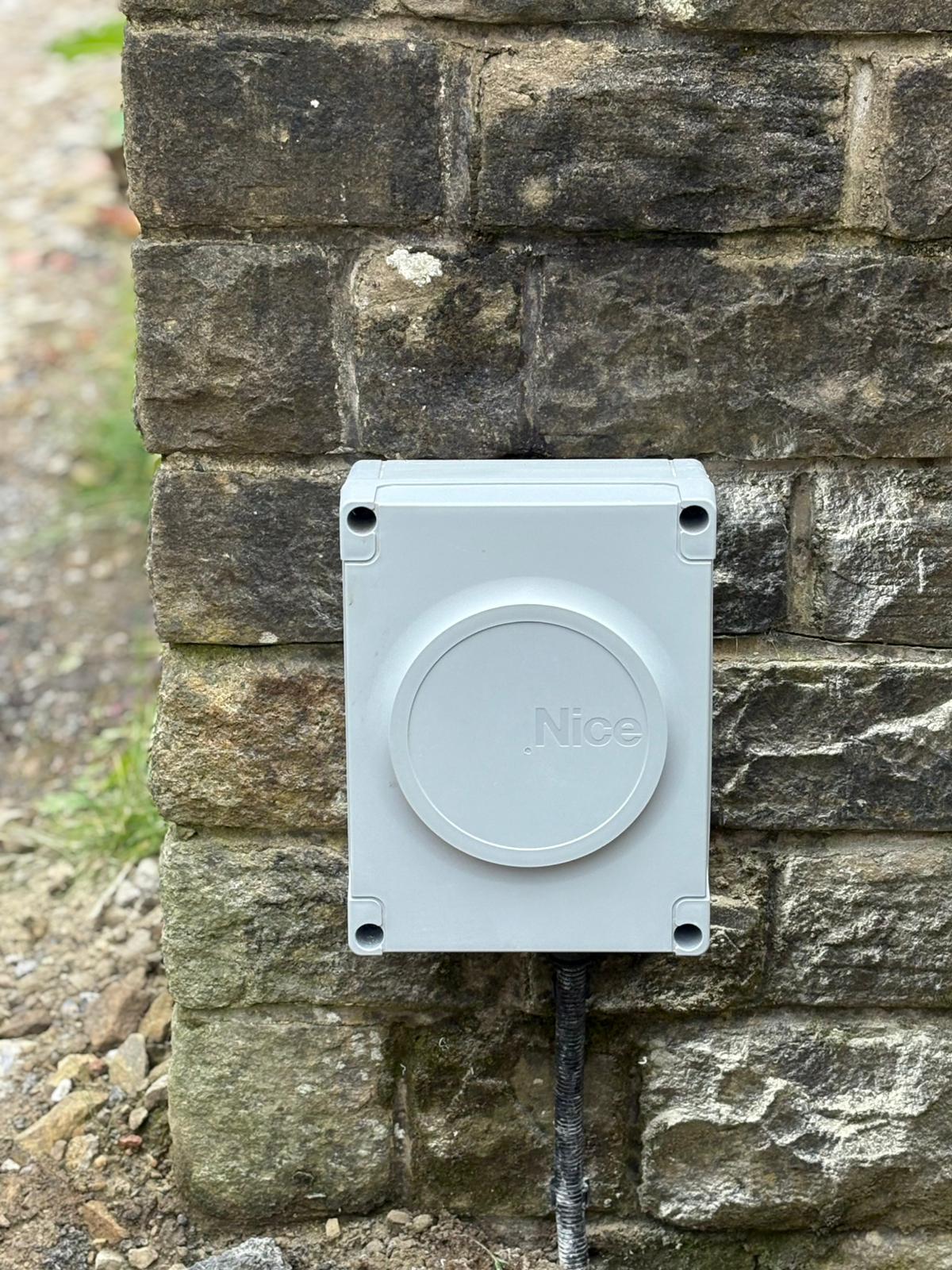 A close-up of a neat, grey Nice branded weatherproof electronic control box mounted flush against a rustic dry-stone wall pillar.