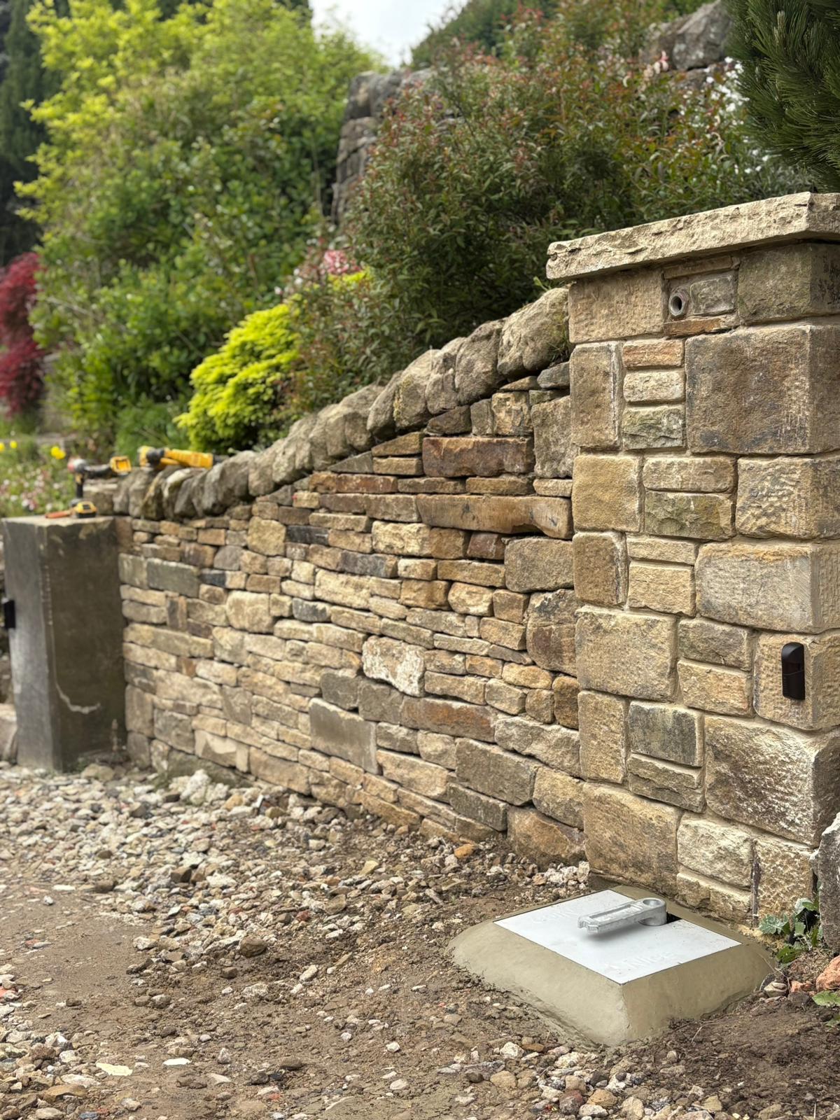 A wide shot of the completed installation showing a beautiful dry-stone wall, an integrated safety photocell sensor, and the top bracket of the Nice underground gate motor neatly finished with a concrete surround.
