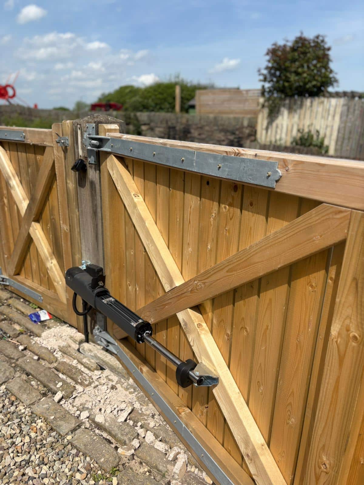 Close-up shot of a light-coloured wooden gate fitted with a sleek black Beninca linear ram automation arm, highlighting the clean installation against the timber framework.