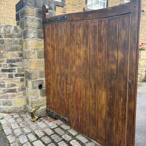 A set of large, dark-stained wooden driveway gates attached to a stone pillar, showing the initial stage of an automation retrofit with a yellow power drill on the cobblestone ground.